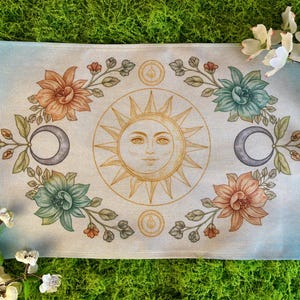 May include: A rectangular, light blue and beige decorative cloth featuring a sun with a face, crescent moons, and floral elements in shades of orange, green, and brown. The cloth is surrounded by green moss and white flowers.