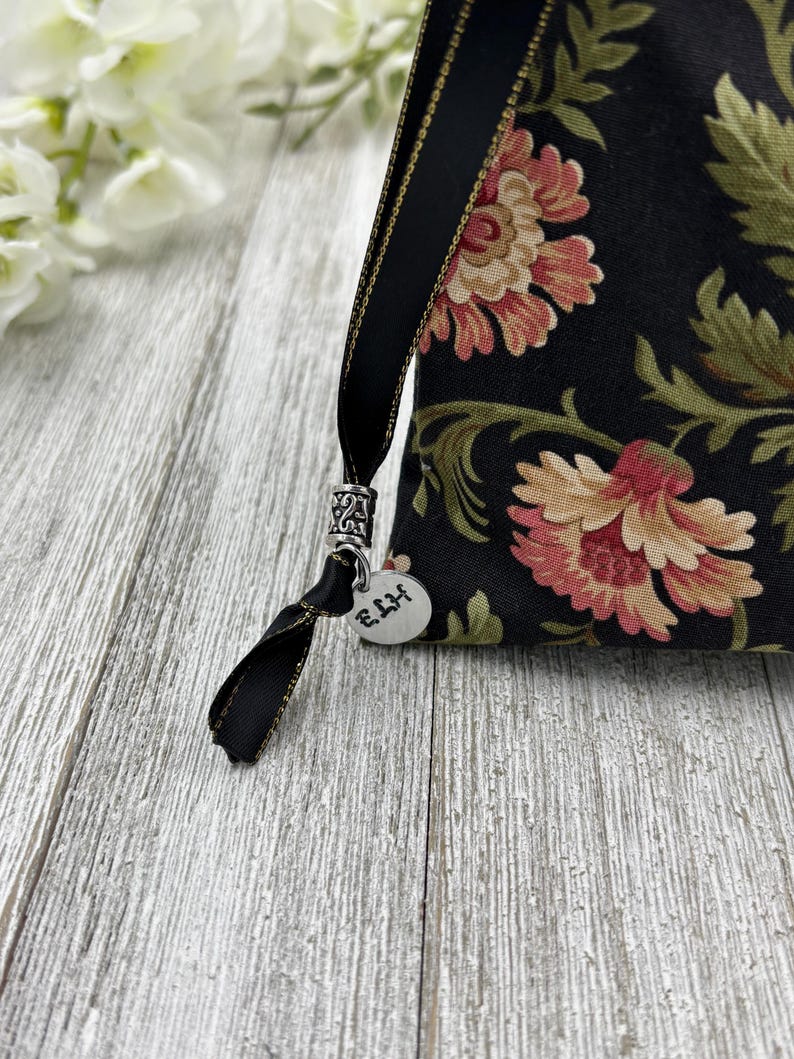 May include: A black fabric pouch with a floral pattern and gold trim. The pouch has a black ribbon with a silver charm that says "E.H.H."
