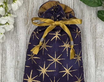 May include: A deep purple fabric drawstring pouch featuring a gold starburst pattern. The bag is cinched at the top with a metallic gold ribbon tied in a bow, accented by two silver-toned cylindrical beads at the ends of the ribbon ties.