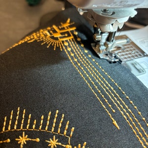 Puede incluir: Primer plano de una m&aacute;quina de coser en funcionamiento, cosiendo hilo dorado sobre tela negra. La tela presenta un dise&ntilde;o bordado con motivos de sol y estrellas, y l&iacute;neas de costura. La aguja est&aacute; en movimiento, creando un patr&oacute;n detallado.