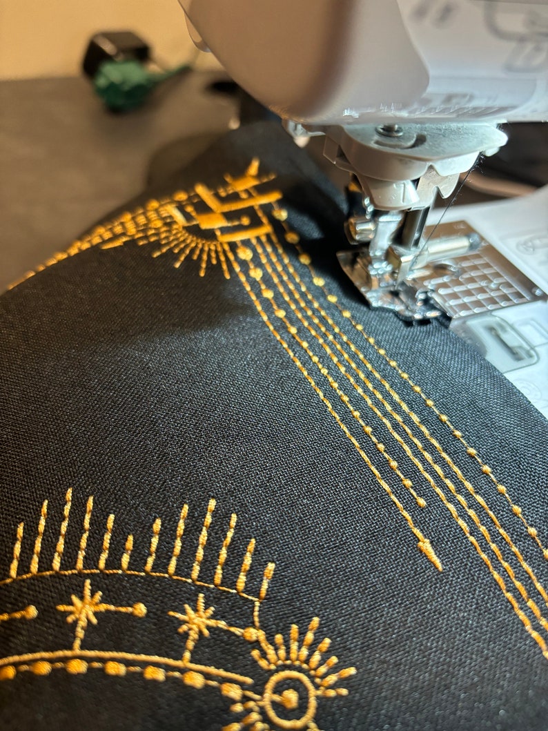 Puede incluir: Primer plano de una m&aacute;quina de coser en funcionamiento, cosiendo hilo dorado sobre tela negra. La tela presenta un dise&ntilde;o bordado con motivos de sol y estrellas, y l&iacute;neas de costura. La aguja est&aacute; en movimiento, creando un patr&oacute;n detallado.
