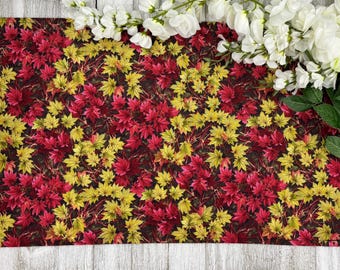 May include: A rectangular fabric with a repeating pattern of red and yellow maple leaves on a dark background. White flowers and green leaves are arranged along the top edge, creating a decorative border.