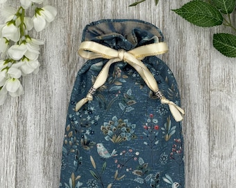 May include: A blue fabric drawstring pouch featuring a repeating pattern of small white rabbits, birds, and various botanical elements. The bag is cinched at the top with a cream-coloured satin ribbon, which is finished with small silver-toned metal beads.