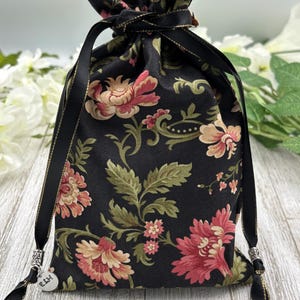May include: Black drawstring bag with a floral pattern and gold trim. The bag has a silver charm with the word "Ellie" on it.