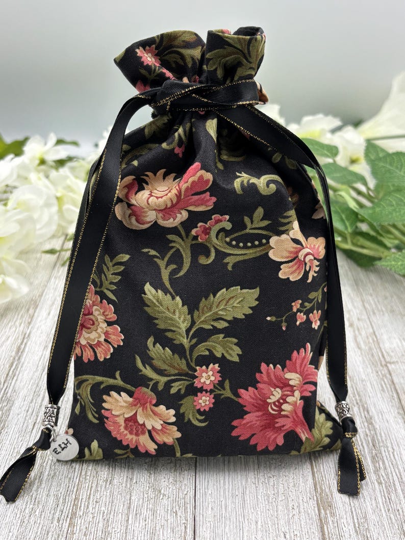 May include: Black drawstring bag with a floral pattern and gold trim. The bag has a silver charm with the word "Ellie" on it.