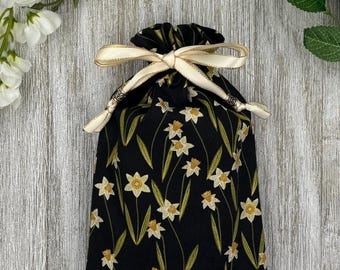 May include: A black drawstring bag with a daffodil pattern. The bag features a cream-colored ribbon with gold trim and decorative silver beads. The daffodils are white and yellow with green stems and leaves.