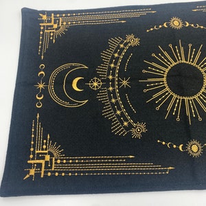Puede incluir: Tela negra con un dise&ntilde;o celestial dorado que presenta una luna creciente, un sol, estrellas y patrones geom&eacute;tricos. El dise&ntilde;o es sim&eacute;trico y cubre la mayor parte de la superficie de la tela. La tela tiene un aspecto ligeramente texturizado.