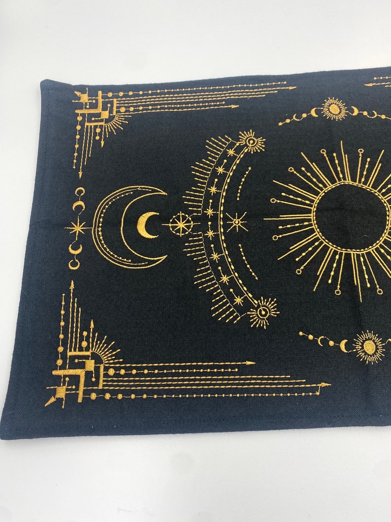 Puede incluir: Tela negra con un dise&ntilde;o celestial dorado que presenta una luna creciente, un sol, estrellas y patrones geom&eacute;tricos. El dise&ntilde;o es sim&eacute;trico y cubre la mayor parte de la superficie de la tela. La tela tiene un aspecto ligeramente texturizado.