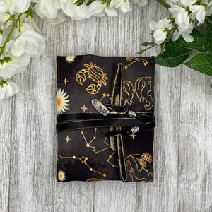 May include: A black fabric journal with gold zodiac constellation designs, wrapped with a black ribbon and a clear crystal charm. The journal is decorated with gold stars and suns. The background is a light wood surface with white flowers.