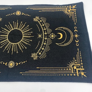 Puede incluir: Textil rectangular negro con dise&ntilde;os celestiales dorados, incluyendo un sol, una luna, estrellas y patrones geom&eacute;tricos. El dise&ntilde;o es sim&eacute;trico, con l&iacute;neas y formas detalladas. El textil se utiliza probablemente con fines decorativos o espirituales.