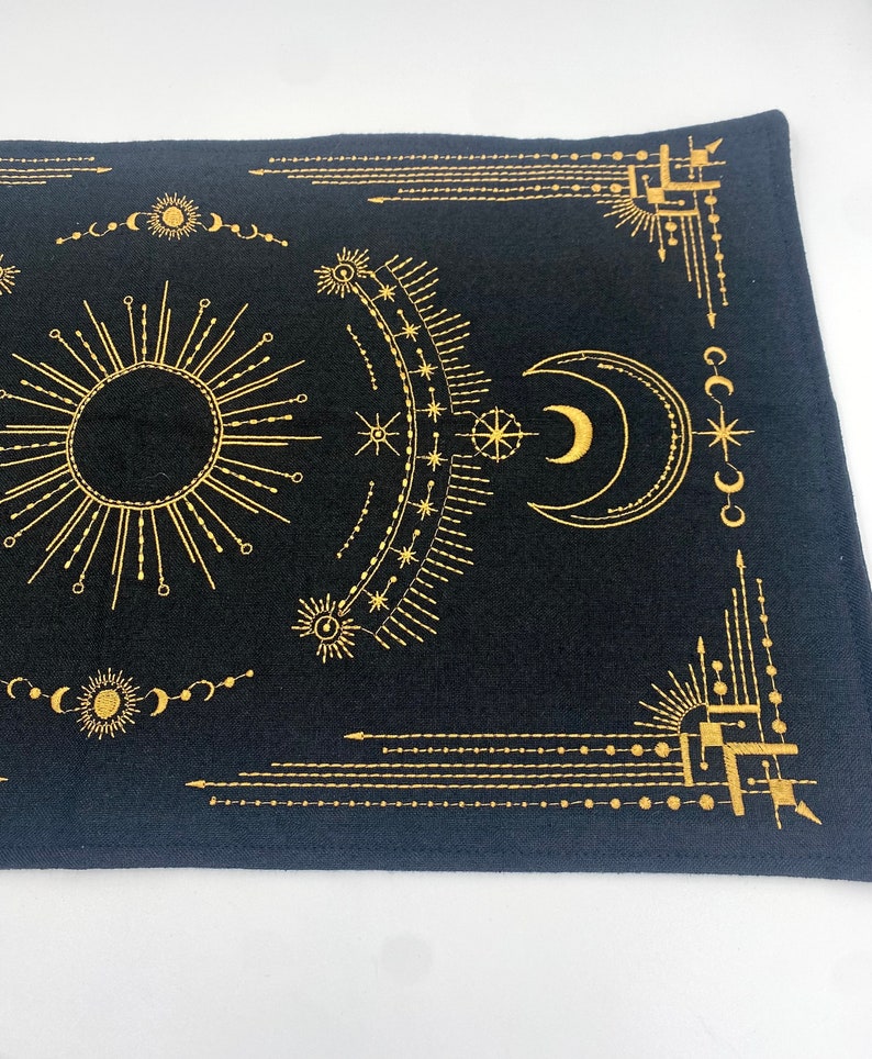 Puede incluir: Textil rectangular negro con dise&ntilde;os celestiales dorados, incluyendo un sol, una luna, estrellas y patrones geom&eacute;tricos. El dise&ntilde;o es sim&eacute;trico, con l&iacute;neas y formas detalladas. El textil se utiliza probablemente con fines decorativos o espirituales.