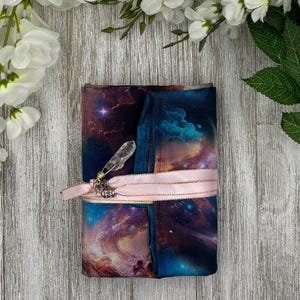May include: A folded fabric item with a celestial galaxy print in shades of blue, purple, and pink. A clear crystal is attached with gold wire. A pink ribbon with gold trim wraps around the item. The item is on a light wood surface with white flowers.