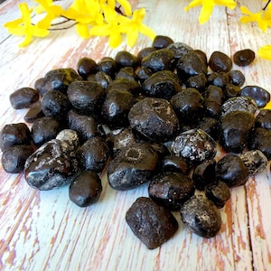 Pode incluir: Uma pilha de pedras pretas polidas com um acabamento brilhante. As pedras variam em tamanho e forma, algumas mostrando veios brancos. Flores amarelas espalhadas no fundo, criando um contraste com as pedras escuras.