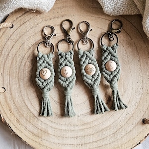 Puede incluir: Cuatro llaveros de macramé verde con cuentas de madera. Cada cuenta tiene un símbolo diferente: un corazón, un ancla, un sol y una luna.