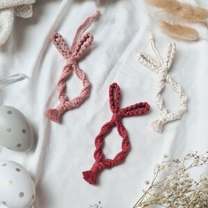 Peut inclure: Trois ornements de lapin en macramé dans les tons de rose et de rouge, avec un blanc, disposés sur un tissu blanc. Des œufs de Pâques décoratifs et des fleurs séchées sont également présents, créant un thème festif et fait main.