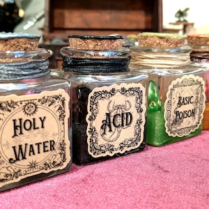 Op de afbeelding: Vier vierkante glazen flessen met kurken stoppen, elk voorzien van een decoratief etiket. De etiketten bevatten de teksten Holy Water, Acid, Basic Poison en Alchemist's Pie. De flessen bevatten verschillende gekleurde vloeistoffen en poeders.