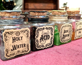 May include: Four square glass bottles with cork stoppers, each featuring a decorative label. The labels read Holy Water, Acid, Basic Poison, and Alchemist's Pie. The bottles contain various colored liquids and powders, wrapped with twine at the neck.