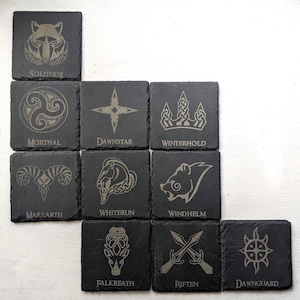 Op de afbeelding: Een verzameling vierkante, donkergrijze leistenen onderzetters. Elke onderzetter heeft een uniek geëtst ontwerp en tekst, waaronder een wolf, een symbool, een ster, een kroon, een ram, een paard, een beer, een hert, gekruiste zwaarden en een zon.