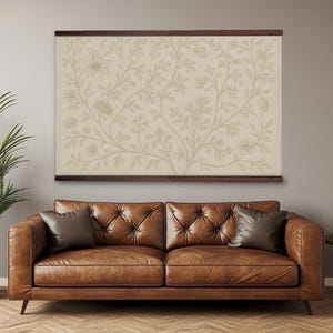 Könnte beinhalten: Ein großes gerahmtes Kunstwerk mit beigem floralem Muster hängt über einem braunen Ledersofa mit zwei Kissen. Das Kunstwerk ist mit dunkelbraunem Holz gerahmt. Eine Topfpflanze befindet sich links.
