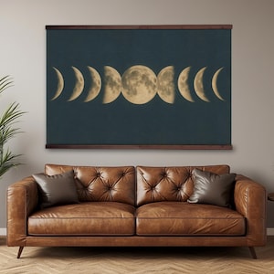 May include: A wall hanging featuring the phases of the moon in shades of gold and cream against a dark teal background. The artwork is framed with a dark brown wooden border and hangs above a brown leather sofa.