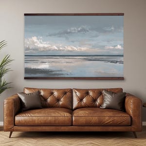 Pode incluir: Uma pintura de paisagem marinha com céu nublado, emoldurada em madeira. A obra de arte está pendurada acima de um sofá de couro marrom com duas almofadas. Uma planta em vaso está à esquerda e uma pequena mesa lateral com uma planta à direita.