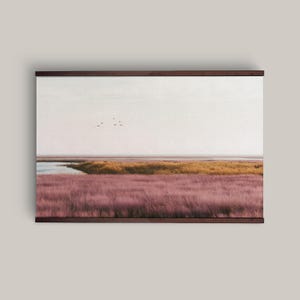 Peut inclure: Une impression murale de paysage horizontal représentant un champ de hautes herbes rose-violet au premier plan, une bande de roseaux jaune doré, un plan d'eau calme et un ciel pâle avec un petit groupe d'oiseaux volant au loin.