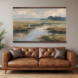 May include: A landscape painting of a marsh scene with a white bird standing in the water. The artwork is framed in a dark brown wooden frame and hangs above a brown leather sofa. The painting features muted colors.
