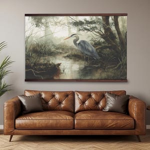 May include: A framed canvas print depicts a serene wetland scene. A great blue heron stands in the water, surrounded by lush greenery and trees. The artwork is mounted on a dark wooden frame, creating a natural and calming aesthetic.