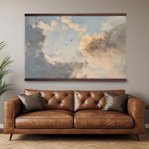 Op de afbeelding: Een groot ingelijst kunstwerk met een bewolkte lucht met twee vogels in vlucht. Het kunstwerk is gemonteerd op een donkerbruin houten frame en hangt boven een gecapitonneerde bruin lederen bank met twee sierkussens. Een potplant staat links.