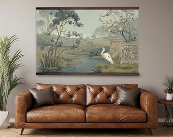 May include: A framed landscape tapestry depicting a serene garden scene with a white heron near a pond. The artwork features muted greens, blues, and browns, and is displayed above a brown leather sofa with two brown pillows.