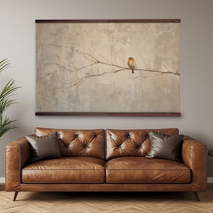 May include: A framed canvas art print featuring a small bird perched on a branch against a textured, neutral-toned background. The artwork hangs above a tufted, brown leather sofa with two throw pillows. A potted plant is to the left.