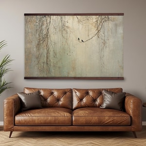 May include: A large rectangular wall art print featuring a serene landscape with two birds perched on a branch. The artwork is framed with a dark brown wooden border. The print hangs above a brown leather sofa with two throw pillows.