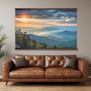 May include: A landscape print of a mountain range at sunset, displayed on a wooden frame. The artwork features a vibrant sky with orange and yellow hues, and the mountains are a mix of blue and green. The print is hanging above a brown leather sofa.