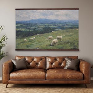 May include: A framed landscape painting depicts sheep grazing in a green pasture with rolling hills and mountains in the background. The artwork is mounted on a wooden frame and hangs above a brown leather sofa.