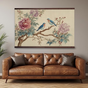 May include: A framed wall art print featuring a watercolor painting of two blue birds perched on a flowering branch. The artwork includes pink peonies, purple flowers, and green leaves against a beige background. The frame is dark brown.