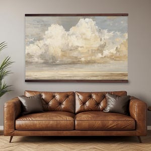 Op de afbeelding: Een groot ingelijst canvasdoek met een bewolkte lucht boven een horizon. Het kunstwerk heeft tinten wit, beige en grijs. Onder het kunstwerk staat een bruine leren bank met twee sierkussens. Links staat een plant in een pot.