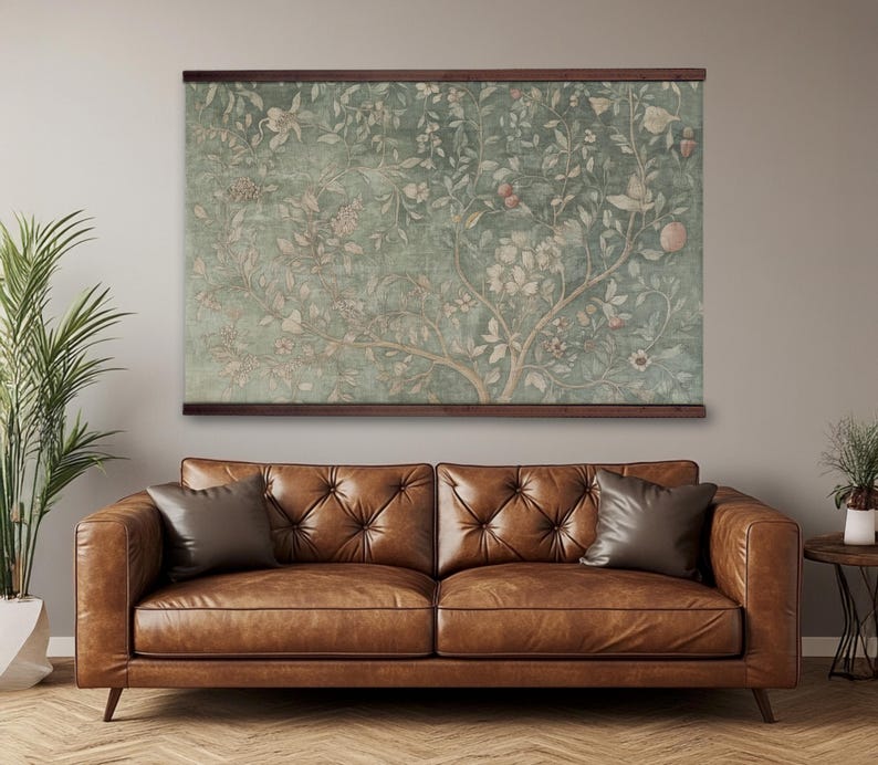 May include: A large wall hanging with a botanical design in shades of green, beige, and pink, framed by a dark brown wooden border. The artwork depicts a tree with flowers and fruit. A brown leather sofa with two pillows is below the artwork.