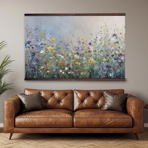 May include: A framed floral painting hangs above a brown leather sofa. The artwork depicts a field of colourful wildflowers in shades of yellow, purple, and white against a soft blue background. The frame is made of wood.