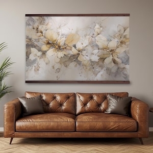 May include: A large floral watercolor print in neutral tones of beige, cream, and gray, mounted on a dark brown wooden frame. The artwork hangs above a brown leather sofa with two throw pillows, creating a warm and inviting living room scene.