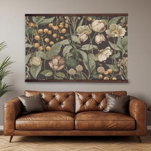 May include: A floral print wall art with a dark brown wooden frame. The artwork features a botanical design with cream, green, and brown flowers and foliage. The artwork hangs above a brown leather sofa with two throw pillows.