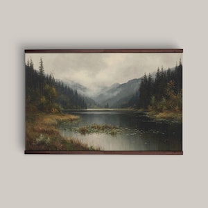 Può includere: Un dipinto di paesaggio incorniciato raffigura un lago sereno circondato da lussureggianti alberi verdi e montagne sotto un cielo nuvoloso. L'opera d'arte è racchiusa in una cornice di legno marrone scuro, aggiungendo un tocco classico alla scena naturale.