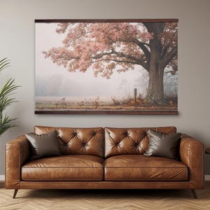 May include: A large framed canvas print depicts a majestic tree with pinkish-red leaves in a misty field. The artwork hangs above a brown leather sofa with two dark brown throw pillows. A potted plant sits to the left, and a small side table is to the right.