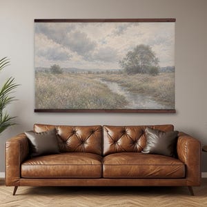 May include: A landscape wall hanging featuring a printed scene of a winding stream through a grassy meadow under a cloudy sky, secured between two horizontal dark wood bars at the top and bottom, displaying muted earth tones and soft blue hues.