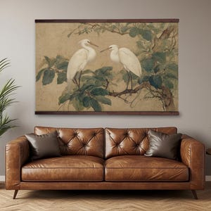 May include: A framed artwork featuring two white birds perched on a tree branch with green leaves. The artwork has a light brown background and is framed in dark brown. The artwork is displayed above a brown leather sofa.