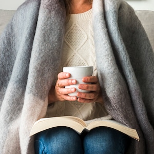 May include: A person is seated, wrapped in a soft, gray and white ombre blanket, holding a white mug. They are wearing a white cable-knit sweater and blue jeans, with an open book in their lap. The scene evokes comfort and relaxation.