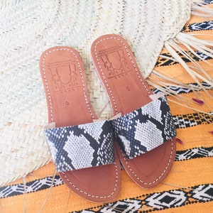 May include: A pair of brown leather sandals with a snakeskin pattern. The sandals have a simple design with a single strap across the top of the foot. The sandals are made by the brand "Artisanat Marocain".