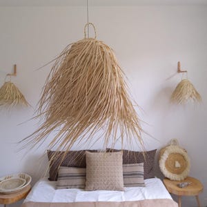 Op de afbeelding: Een slaapkamer met een wit bed, bruine kussens en een grote geweven hanglamp boven het bed. Er zijn twee kleinere geweven hanglampen aan de muren. De kamer heeft een bohemian stijl.