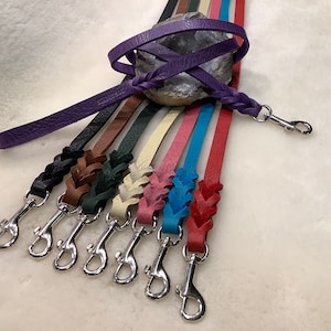 Op de afbeelding: Een set van acht leren hondenriemen in verschillende kleuren, waaronder paars, bruin, groen, crème, roze, blauw en rood. Elke riem heeft een gevlochten ontwerp en een zilveren haak.