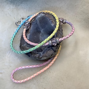 May include: A rainbow-colored braided leather leash with a silver clasp and decorative beads. The leash is wrapped around a dark gray stone, showcasing a gradient of pastel colors including pink, yellow, green, and blue.