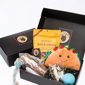 May include: A black gift box containing a yellow bag of Bocce's Soft & Chewy peanut butter and banana treats, a plush taco toy, a rope toy with orange and blue threads, a llama-shaped cookie, and a small light blue ball on black paper filler.